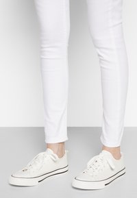 White canvas sneakers feature a textured sole, flat laces, and a contrasting black stripe along the bottom. Worn with white slim-fit jeans.