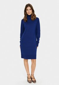Abito a maglia blu con collo alto, maniche lunghe e vestibilità comoda, che arriva a metà coscia; abbinato a tacchi a strappo con stampa leopardata.