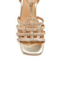 Sandalia dorada de punta cuadrada con tiras trenzadas. Presenta un tacón plano y un acabado metálico suave, proporcionando un diseño abierto y transpirable.