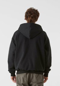 Textúrált anyagból készült fekete kapucnis pulóver, nagy kapucnival, ejtett vállakkal és rugalmas mandzsettákkal és alsó szegéllyel. Cargo nadrággal viselve.