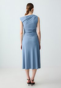 Abito monospalla in tessuto azzurro chiaro, corpino aderente con drappeggio, gonna fluida che arriva a metà polpaccio, abbinato a sandali neri con tacco.
