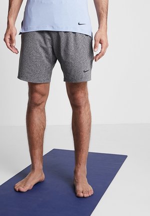 Mężczyzna stojący boso na niebieskiej macie do jogi, ubrany w szare spodenki Nike i jasnoniebieską koszulkę na jednolitym tle.