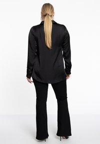 Zwarte satijnen blouse met lange mouwen en een puntige kraag, met zijsplitten en een comfortabele pasvorm, gecombineerd met zwarte flared broeken en hakken.