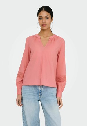 Femme portant des boucles d'oreilles créoles, une blouse corail à manches longues avec des détails en crochet sur les manches, et un jean taille haute bleu clair.