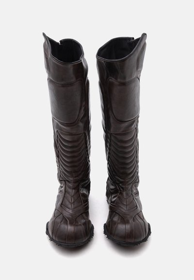 Paire de bottes hautes en cuir marron foncé avec zones rembourrées au niveau des genoux et des tibias, détails côtelés et semelles robustes, debout sur un fond blanc.