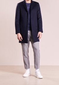 Hombre vestido con un abrigo azul marino sobre un suéter azul marino, pantalones grises a medida, calcetines blancos y zapatillas blancas, de pie contra un fondo beige.