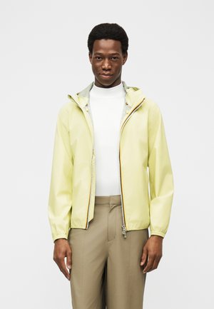 Νέος άνδρας που φοράει ανοιχτό κίτρινο φούτερ με φερμουάρ και κουκούλα, λευκό ζιβάγκο και μπεζ παντελόνι, στέκεται μπροστά σε ένα απλό φόντο.