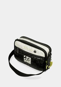 Bolso rectangular en blanco y negro con cremalleras dobles, una correa de tela y un parche con el logo; presenta un patrón de texto y un acento verde en la cremallera.