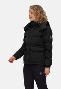 Svart puffjacka med huva, stora framfickor och knäppning. Har ett texturerat tyg och en distinkt logotyp på ärmen.