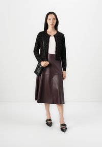 Cardigan noir à motif, haut rose clair, jupe en cuir bordeaux, chaussures noires à talons pointus avec des lanières, et un petit sac à main noir.