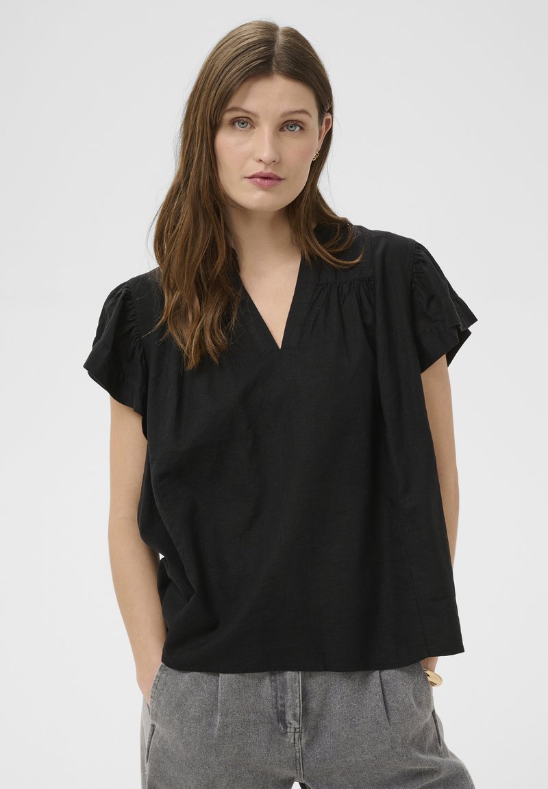 Jeune femme aux longs cheveux bruns portant un haut noir ample à manches courtes et un pantalon gris, debout les mains dans les poches devant un fond uni.