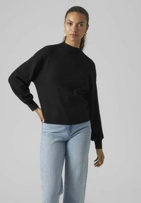 Maglione nero lavorato a maglia con collo alto, vestibilità rilassata e maniche lunghe, abbinato a jeans a gamba larga blu chiaro. Texture liscia.
