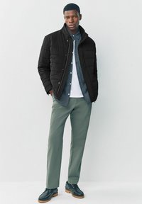Sort quiltet jakke, grønne bukser, hvid skjorte og rutet skjorte med knapper. Sorte sko med en brun sål. Neutral baggrund.