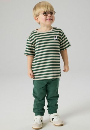 Jonge jongen met blond haar, ronde bril, groen en beige gestreept shirt met voetbalpatch, groene broek en witte sneakers, glimlachend.