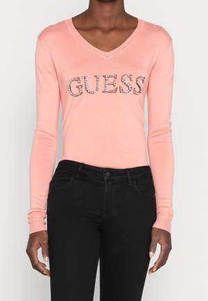 Kobieta w różowym swetrze z dekoltem w serek z literami "GUESS" wykonanymi z kryształków, zestawionym z czarnymi spodniami z wysokim stanem, widoczna od szyi do bioder.