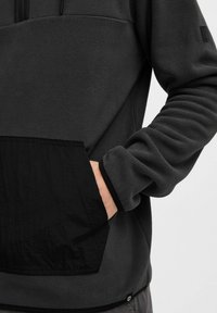 Fleece pullover in donkergrijs met een zwarte zak. Heeft een ritshals, zachte textuur en elastische manchetten. Minimale branding op de zoom.