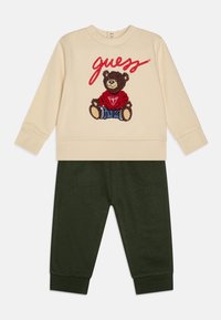 Κρεμ φούτερ με σχέδιο αρκούδας και "guess" σε κόκκινα γράμματα, συνδυασμένο με σκούρα πράσινα παντελόνια, και τα δύο κατασκευασμένα από μαλακό ύφασμα.