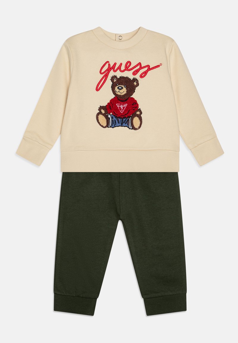 Κρεμ φούτερ με σχέδιο αρκούδας και "guess" σε κόκκινα γράμματα, συνδυασμένο με σκούρα πράσινα παντελόνια, και τα δύο κατασκευασμένα από μαλακό ύφασμα.