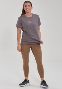Grå atletisk t-shirt med teksten "ENDURANCE", kombineret med brune leggings og sorte sportssko. Enkel design, tætsiddende form, glat tekstur.