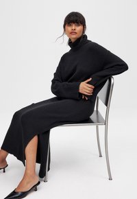 Lungo vestito nero a maglia con colletto alto e spacchi laterali, tessuto testurizzato, abbinato a mule nere con tacco, seduta su una sedia argentata.