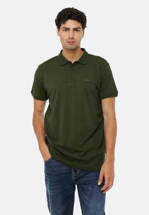 Uomo in piedi con le mani nelle tasche che indossa una polo verde scuro e jeans blu su sfondo bianco.