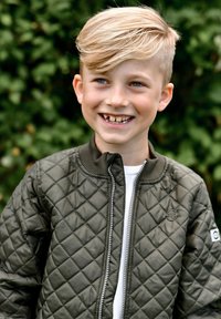 Prešívaná bombera olivovo zelenej farby so zipsom na prednej strane, rebrovaným golierom a logom. Nosí sa cez biele tričko. V pozadí je zeleň.