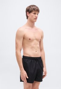 Μαύρα μαγιό σορτς με ελαστική μέση και κορδόνι. Το ύφασμα έχει λεία, γυαλιστερή υφή με απλό σχέδιο.