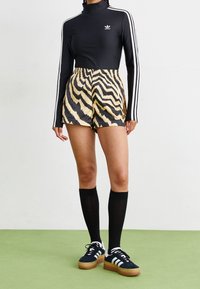 Person i svart Adidas långärmad tröja med vita ränder, beige och svart djurmönstrade shorts, svarta knästrumpor och svarta sneakers.