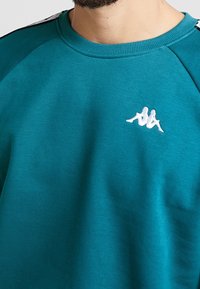 Detailní záběr na tyrkysovou mikinu s kulatým výstřihem, která na hrudi nese bílé logo dvou sedících postav zády k sobě.