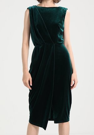 Bez rękawów, ciemnozielona aksamitna sukienka midi z asymetryczną spódnicą kopertową i dopasowaną talią na stojącej kobiecie.