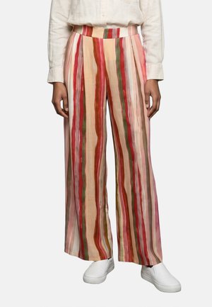 Persoon draagt wijde broek met verticale rode, roze, beige en groene strepen, gecombineerd met een wit overhemdbloesje en witte instapschoenen.