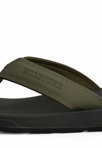 Zbliżenie na oliwkowozielony skórzany pasek sandała z wytłoczonym napisem "HILFIGER" na teksturowanej czarnej podeszwie.
