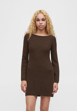 Jeune femme aux longs cheveux blonds bouclés portant une robe moulante à manches longues en tricot côtelé marron foncé, debout devant un fond uni.