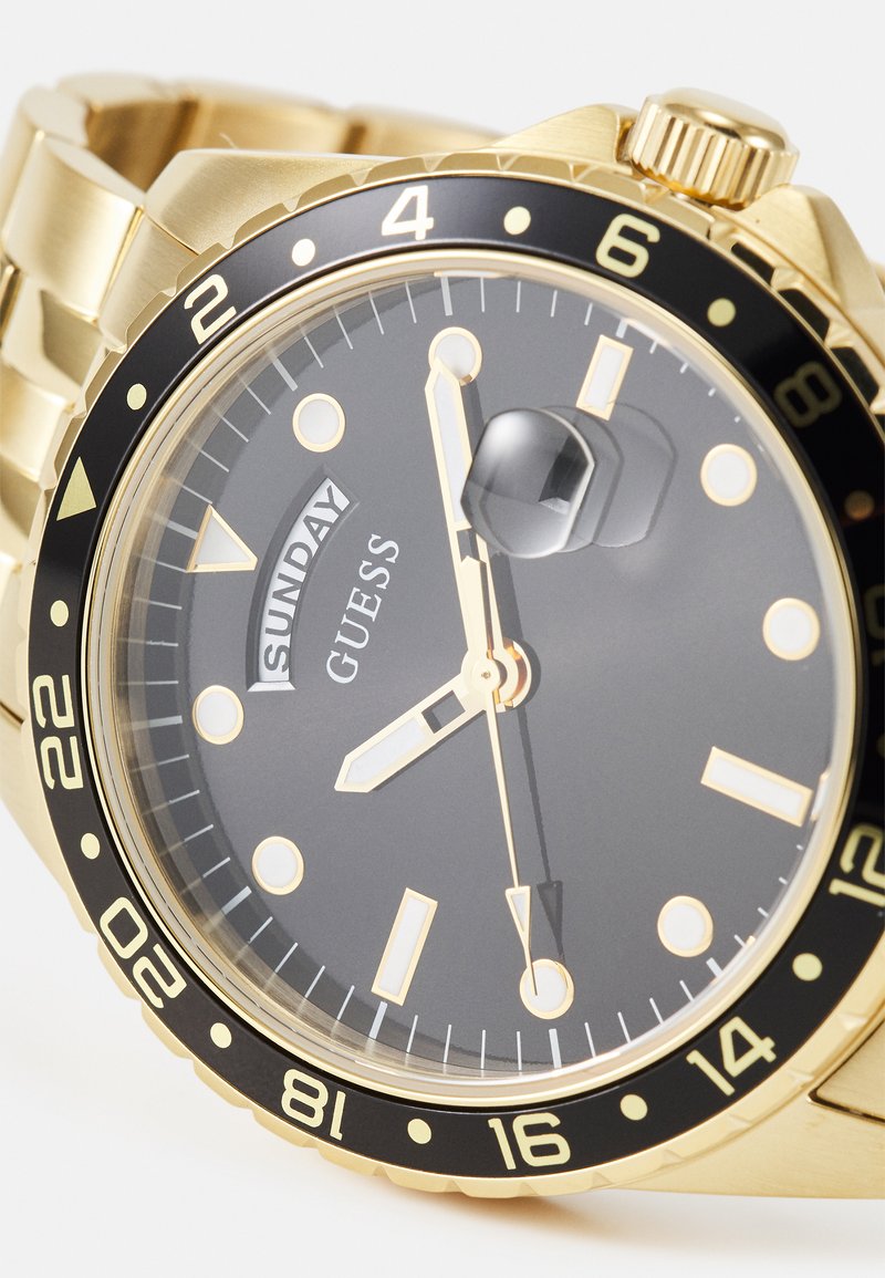 Reloj de acero inoxidable dorado con una esfera negra, marcadores de hora dorados y un bisel negro con números blancos. Indicador del día a las 12 en punto.