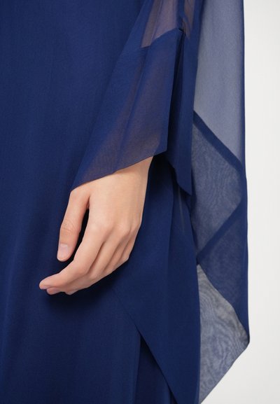 Rochie bleu marin cu mânecă din chiffon fluid și design stratificat al textilei. Mâna prezentată este relaxată și poziționată aproape de marginea țesăturii.