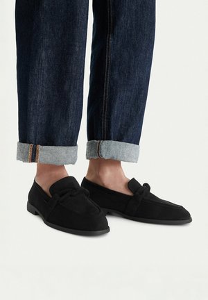 Persona con vaqueros oscuros remangados y mocasines de ante negros con detalle de lazo, de pie sobre una superficie clara.