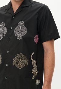 Camisa negra de manga corta con intrincados bordados multicolores de patrones florales y un tigre, con un cierre de botones en el frente y un corte relajado.
