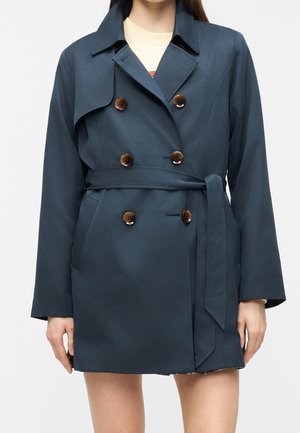 Kvinde iført en mørkeblå dobbeltradet trenchcoat med brune knapper og et bundet bælte i taljen over en lysefarvet top.