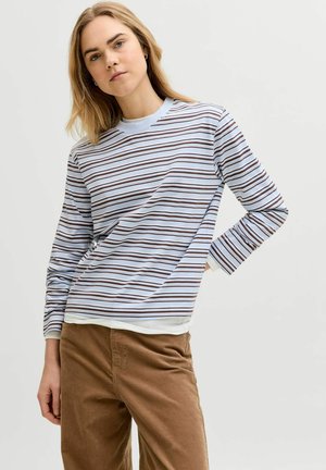 Blondide juustega naine, kes kannab helesinist ja pruunide triipudega pika varrukaga särk ja pruune pükse, poseerides ühe käega puusal.