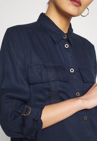 Marineblaues Button-Up-Hemd mit zwei Brusttaschen, hochgekrempelten Ärmeln, die mit einem Metallring gesichert sind, glattes Gewebe mit einem subtilen Glanz.
