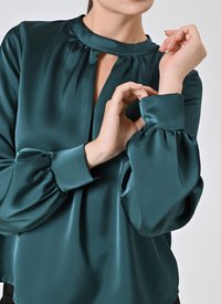 Blusa di raso teal con collo alto, dettaglio a chiave e maniche a sbuffo. Texture liscia, design fluido e polsini aderenti.