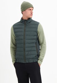 Groene gewatteerde vest met hoge kraag en ritsluiting, gedragen over een gestructureerde groene trui. Model draagt een bijpassende gebreide muts.