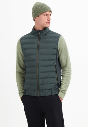 Grüne gepolsterte Weste mit hohem Kragen und Reißverschluss, über einem strukturierten grünen Pullover getragen. Model trägt eine passende Strickmütze.