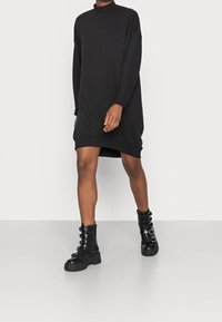 Černé oversize šaty ve stylu svetru s vysokým límcem, dlouhými rukávy a žebrovanými manžetami, doplněné černými šněrovacími bohatýrskými botami.