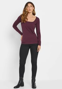 Pitkähihainen, pyöreäkauluksinen toppi syvässä purppuravärissä; istuva muotoilu sileällä tekstuurilla, yhdistetty mustiin skinny-farkkuihin ja nilkkureihin.