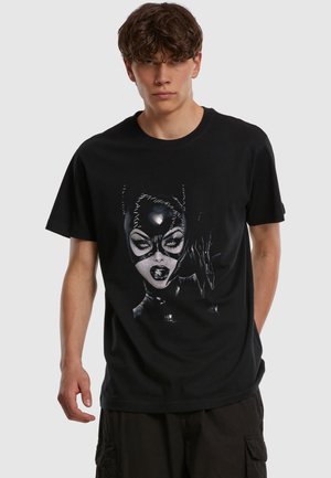 Joven con una camiseta negra con un gráfico detallado de una mujer con una máscara de gato de cuero cosida y guantes sobre un fondo sencillo.