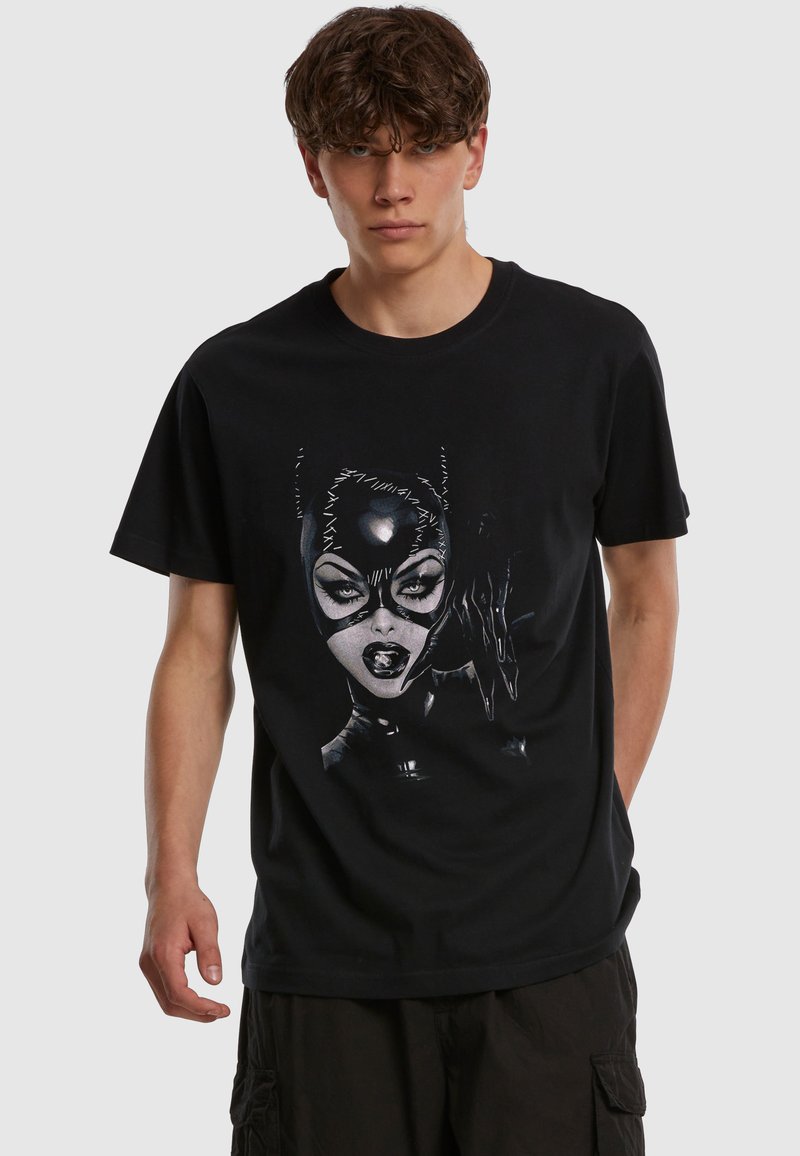 Νέος άνδρας που φοράει μαύρο T-shirt με λεπτομερή γραφικά μιας γυναίκας με ραμμένη δερμάτινη μάσκα γάτας και γάντια σε μονόχρωμο φόντο.