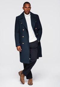 Dunkelblauer Wollmantel mit doppelreihigem Design und goldenen Knöpfen, getragen über einem weißen Strickpullover mit Rollkragen und schwarzen Hosen. Braune Stiefeletten.