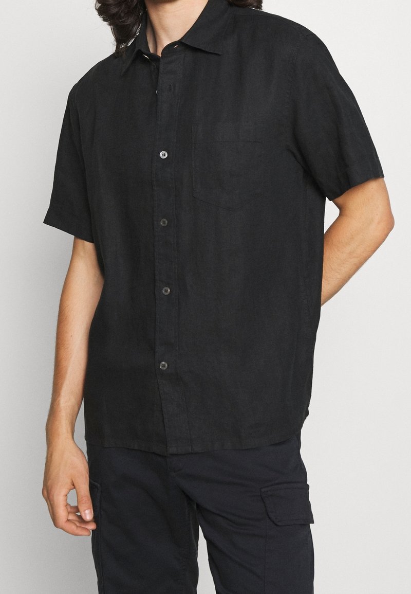 Homme portant une chemise noire à manches courtes avec une poche poitrine et un pantalon cargo noir sur un fond uni.