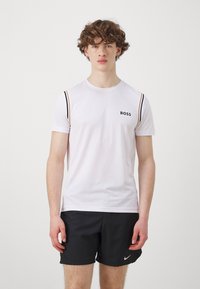 Camiseta atlética de manga corta blanca con el logo negro "BOSS", que presenta rayas horizontales tonales en las mangas, combinada con pantalones cortos negros.
